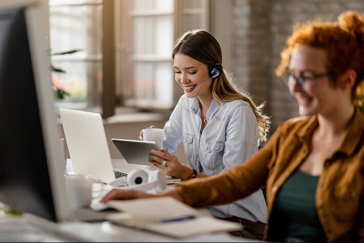 Happy female entrepreneur with headset drinking coffee while surfing the net on touchpad in the office